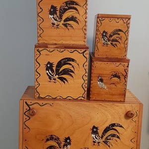 Può includere: Una collezione di scatole di legno e un schedario, tutti con disegni di galli dipinti a mano. Le scatole variano in dimensioni e sono impilate. L'opera d'arte del gallo è in bianco e nero con accenti gialli. Le scatole hanno una finitura marrone chiaro.