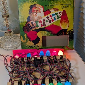 May include: A vintage Noma Santa Lites 15-light set in a green cardboard box featuring a Santa Claus illustration. The box text reads 15 LIGHT SET, MULTIPLE WIRING, FOR INDOOR USE, 110-120 VOLTS, EACH LAMP BURNS INDEPENDENTLY. The string of lights is lit.
