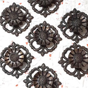 Puede incluir: Siete tiradores de gabinete de color bronce con un diseño floral decorativo. Cada tirador presenta un centro circular con líneas radiales y está enmarcado en un marco ornamental y simétrico. Herrajes para muebles.