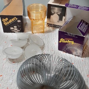 May include: A collection of vintage toys including a Slinky, a set of floating candles, and a square egg maker. The Slinky is silver and coiled. The candles are white and in clear plastic containers. The egg maker is clear plastic and has a white egg inside.