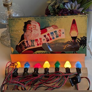 May include: A vintage Noma Santa Lites eight-light set in a rectangular cardboard box. The box features a graphic of Santa Claus and the text SANTA LITES. The set includes seven colourful light bulbs in yellow, blue, pink, and green, connected by red and green electrical wires, resting in a cardboard tray.