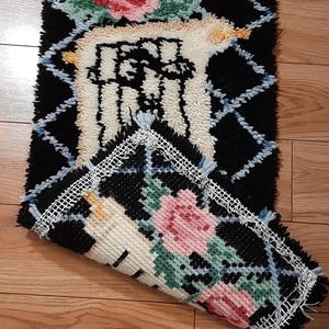 Puede incluir: Una alfombra rectangular con un patrón de diamantes negros y detalles florales. La alfombra presenta rosas rosas y hojas verdes. Los bordes están adornados con un borde blanco similar al encaje. Parte de la alfombra está enrollada, revelando la parte inferior.