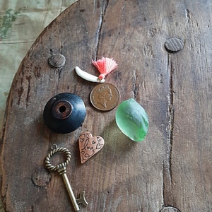 Puede incluir: Una superficie de madera con una variedad de objetos pequeños, incluyendo una llave, un amuleto en forma de corazón con la palabra "love" inscrita en él, una pequeña pieza de vidrio verde, una moneda con el perfil de una mujer y un diente blanco con una borla roja.