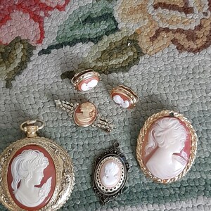 May include: A collection of six cameo jewellery pieces featuring portraits of women. The pieces include a watch, a pendant, a brooch, and two pairs of earrings. The cameos are set in gold-tone metal and feature white cameo portraits against a red background.