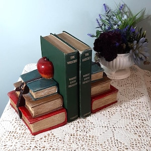 May include: Two green hardback books titled "The Wrecker" by Robert Louis Stevenson, with decorative bookends. The bookends are designed as stacks of books in red, brown, and blue, with a red apple on top. A vase of flowers is in the background.