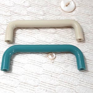 Peut inclure: Deux poignées de meuble, une crème et une turquoise, sont présentées. Chaque poignée est de forme rectangulaire incurvée avec des trous à chaque extrémité. Deux boutons, un blanc et un nacré, sont également visibles.