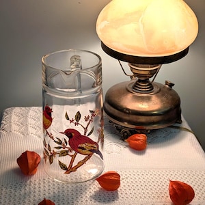 Könnte beinhalten: Eine klare Glaskaraffe mit Henkel, verziert mit roten und gelben Vögeln auf Ästen. Neben der Karaffe steht eine antike Messinglampe mit cremefarbenem Schirm. Orangefarbene chinesische Lampionblumen sind verstreut.