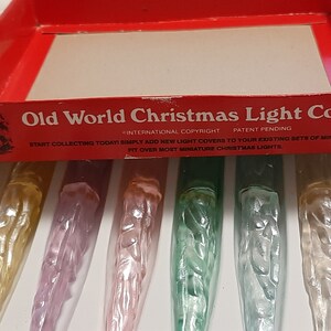 May include: A box of Old World Christmas Light Covers. The box is red with white lettering and a picture of Santa Claus. The box contains six light covers, each with a different design. The light covers are made of clear plastic and have a frosted finish. The light covers are designed to fit over miniature Christmas lights.