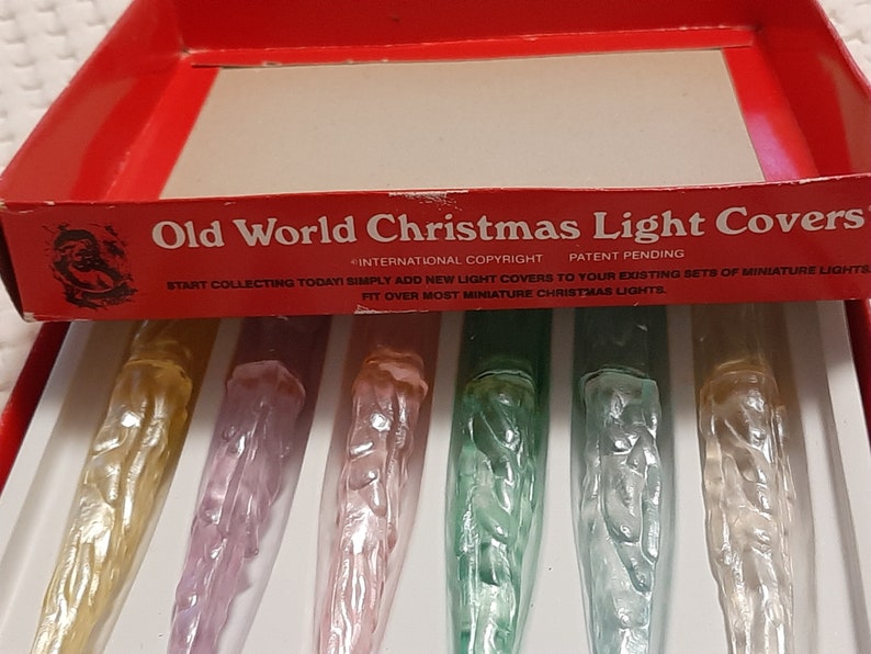 May include: A box of Old World Christmas Light Covers. The box is red with white lettering and a picture of Santa Claus. The box contains six light covers, each with a different design. The light covers are made of clear plastic and have a frosted finish. The light covers are designed to fit over miniature Christmas lights.
