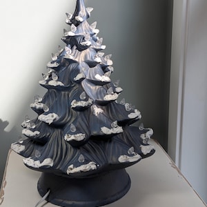 May include: A ceramic tabletop Christmas tree lamp in a dark navy blue finish with a wood-grain texture. The tree features white painted snow accents on the branches and numerous clear, teardrop-shaped plastic bulbs inserted into the surface.