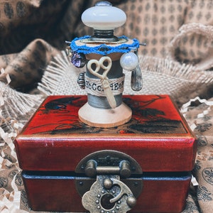 May include: A vintage wooden spool with a white ceramic knob, blue thread, and a pair of scissors, sitting on a red lacquered box with a brass latch. The spool is decorated with the text "BbCcEeF".