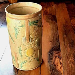 Puede incluir: Un jarrón cilíndrico de cerámica alta con una base de color beige. El jarrón presenta diseños en relieve de sol y hojas en amarillo y verde. Dos asas verticales están en los lados. El jarrón está sobre una superficie de madera.