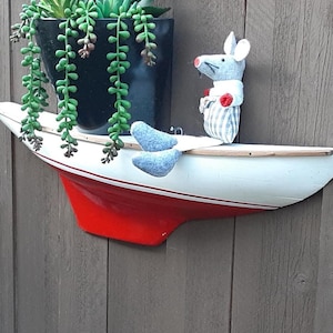 May include: A decorative sailboat-shaped shelf with a red hull and white deck. A small, grey mouse figure with a red bow tie and striped trousers stands on the deck. A black pot with green plants is mounted above the shelf.
