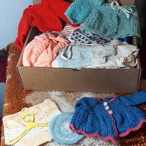 Puede incluir: Una caja de cartón llena de ropa infantil vintage. Los artículos incluyen un suéter rojo tejido, una blusa de ganchillo azul claro con un lazo blanco y una prenda de punto color melocotón. También hay una chaqueta de punto azul y rosa y una percha amarilla.