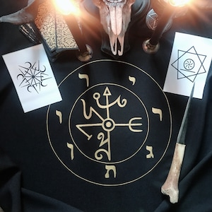 Pode incluir: Um pano preto com um círculo dourado contendo uma bússola e letras hebraicas. Há duas velas, uma caveira e dois cartões com símbolos neles.