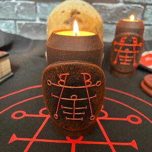 May include: Two brown candles with red sigils on the sides, one lit and one unlit, sitting on a black surface with a red circle and lines.