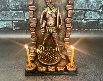 May include: A bronze-toned sculpture of a horned figure with red eyes, standing within a stone-textured frame. The figure wears a necklace and belt, with a tail and cloven hooves. A circular sigil rests at its feet, flanked by two thin yellow candles.