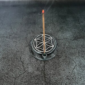 May include: A brown incense stick burning in a black circular incense holder with a white geometric design. The holder is on a gray surface.