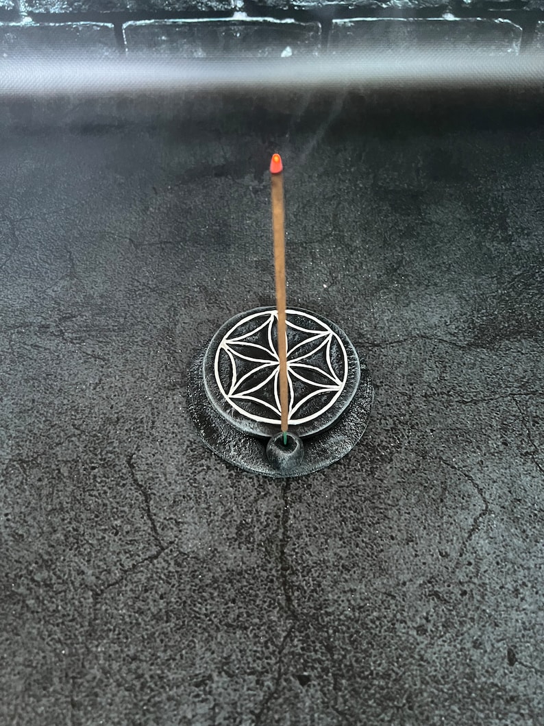 May include: A brown incense stick burning in a black circular incense holder with a white geometric design. The holder is on a gray surface.