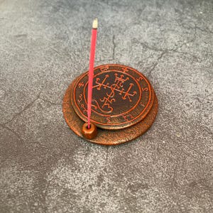 May include: A bronze-colored incense holder with a circular design and a red incense stick. The holder has a layered design with a detailed pattern in a darker shade of bronze. The incense stick is red with a white tip.