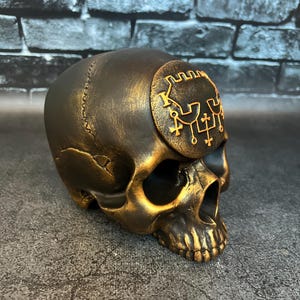 Könnte beinhalten: Eine dekorative Totenkopfskulptur mit dunkler Bronzefinish und goldenen Akzenten. Der Schädel weist ein detailliertes Design auf, einschließlich eines kreisförmigen Emblems mit komplizierten Symbolen auf der Stirn. Die Skulptur steht auf einer strukturierten Oberfläche.