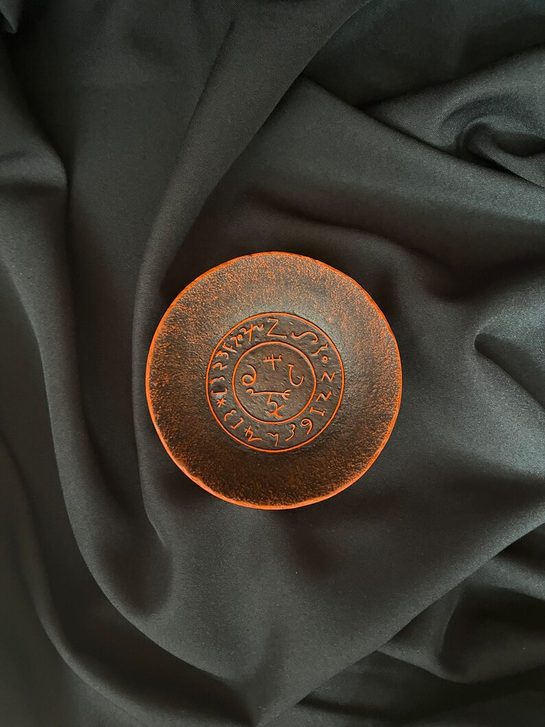 K&ouml;nnte beinhalten: Eine kleine, runde Schale aus terracotta in Terrakottafarbe mit einem erhabenen, kreisf&ouml;rmigen Design in der Mitte. Das Design zeigt ein Symbol mit einem Schl&uuml;ssel und anderen Markierungen, umgeben von Zahlen.