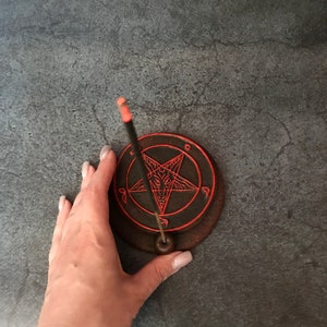 May include: A black and red ceramic incense holder with a pentagram design. A black incense stick is placed in the holder.