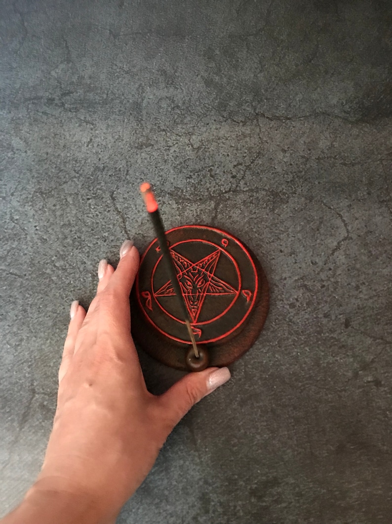 May include: A black and red ceramic incense holder with a pentagram design. A black incense stick is placed in the holder.
