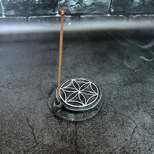 May include: A gray ceramic incense holder with a white geometric design. A brown incense stick is burning in the holder.