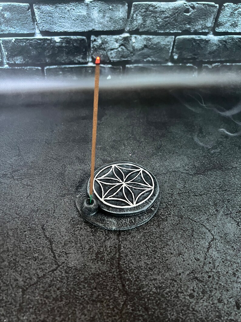 May include: A gray ceramic incense holder with a white geometric design. A brown incense stick is burning in the holder.
