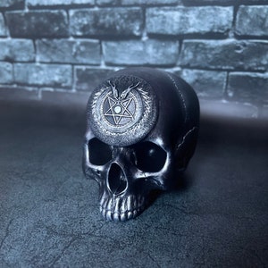 Könnte beinhalten: Eine schwarze Schädel-Figur mit einem silbernen Pentagramm-Emblem auf der Oberseite. Der Schädel hat detaillierte Merkmale und sitzt auf einer dunklen Oberfläche. Das Emblem ist von einem kreisförmigen Design umgeben.