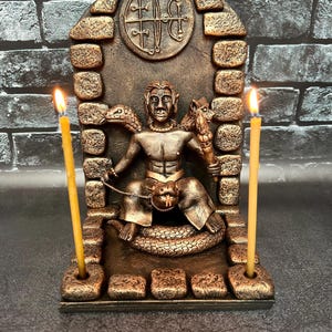 May include: A bronze-coloured statue of a deity with snakes and a wolf, sitting on a throne with a symbol above. Two lit candles are on either side of the statue.