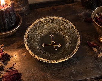 May include: A small, round, dark-colored bowl with a textured, uneven surface and a metallic gold rim. The interior of the bowl features a centered, engraved gold symbol consisting of a vertical line with a crossbar and three small crosses.