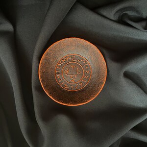 K&ouml;nnte beinhalten: Eine kleine, runde, orange-braune Keramikschale mit einem schwarzen Kreisdesign und Symbolen in der Mitte. Die Schale befindet sich auf einem schwarzen Stoffhintergrund.