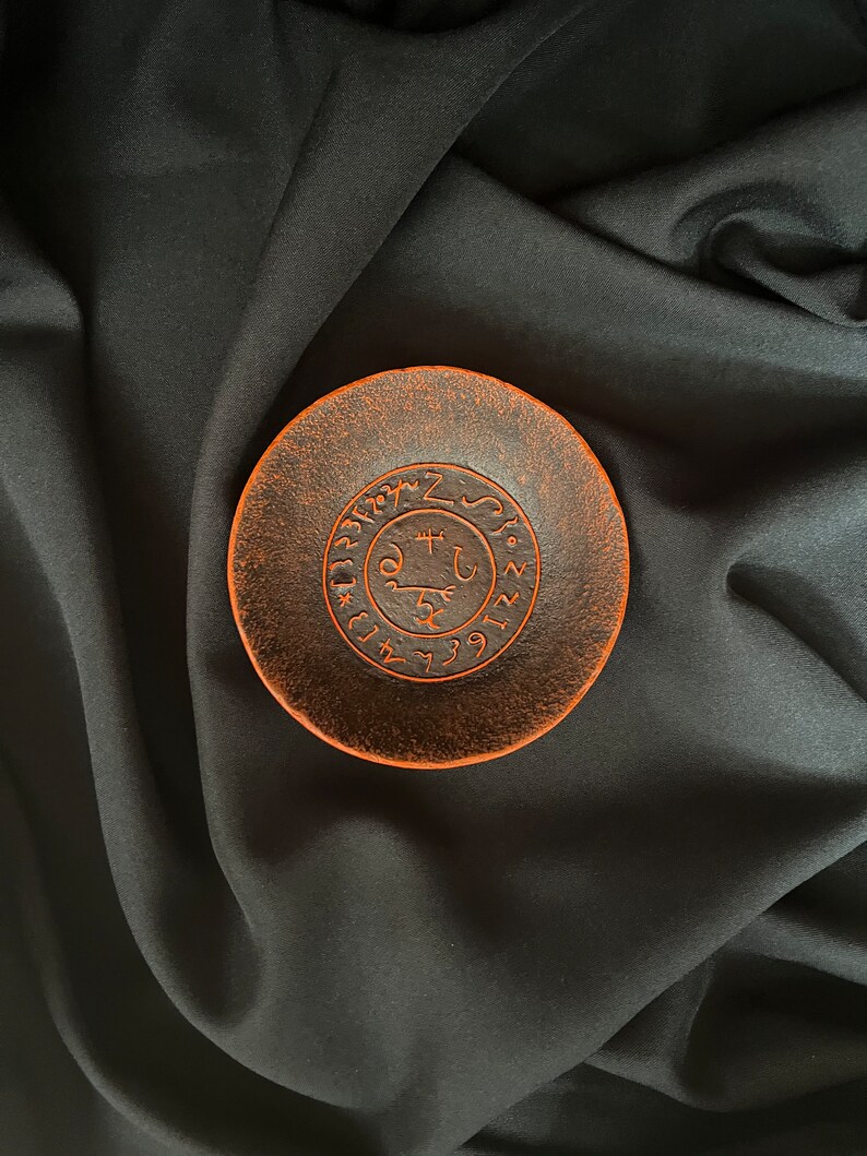 K&ouml;nnte beinhalten: Eine kleine, runde, orange-braune Keramikschale mit einem schwarzen Kreisdesign und Symbolen in der Mitte. Die Schale befindet sich auf einem schwarzen Stoffhintergrund.