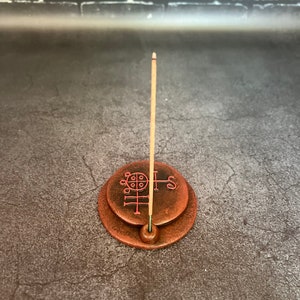 Könnte beinhalten: Ein brauner Keramik-Räucherstäbchenhalter mit einem roten Sigel-Design. Ein Holz-Räucherstäbchen ist in den Halter gelegt.