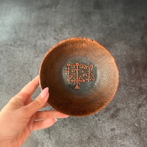May include: A small, round, brown ceramic dish with a black interior. The dish has a red sigil or symbol in the center.