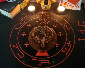 May include: A black felt mat with a red circle containing a pentagram and other symbols. A skull with a pentagram on its forehead sits on the mat. A deck of tarot cards and two candles are also on the mat.