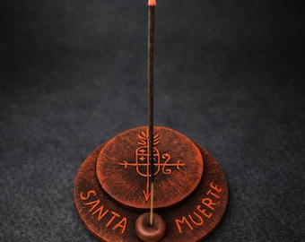 Può includere: Un porta incenso rotondo marrone scuro dal design a gradini, caratterizzato dalla scritta arancione "SANTA MUERTE" e da un simbolo centrale raffigurante un ananas con una freccia. Un bastoncino d'incenso è inserito ed emette del fumo.