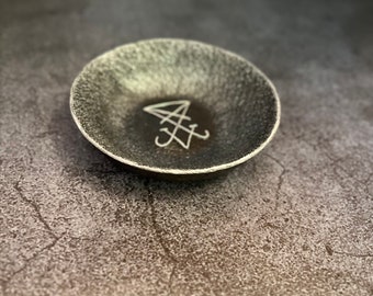 May include: A small, round, dark gray ceramic bowl with a textured, speckled exterior and a smooth interior. The center of the bowl features a white, hand-drawn sigil consisting of intersecting lines and geometric shapes.