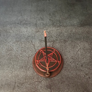 May include: A black incense stick burning on a brown ceramic incense holder with a red pentagram design.
