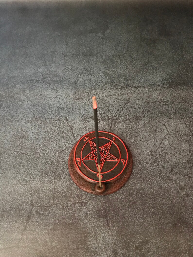 May include: A black incense stick burning on a brown ceramic incense holder with a red pentagram design.