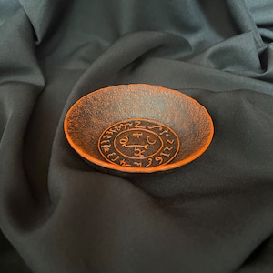 K&ouml;nnte beinhalten: Eine schwarze Metallschale mit einem orangefarbenen Design eines Kreises mit Symbolen und Zahlen darin. Die Schale befindet sich auf einem schwarzen Stoffhintergrund.