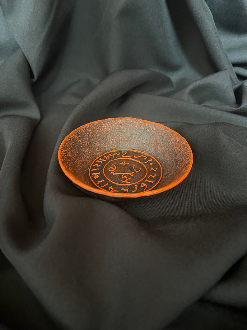 K&ouml;nnte beinhalten: Eine schwarze Metallschale mit einem orangefarbenen Design eines Kreises mit Symbolen und Zahlen darin. Die Schale befindet sich auf einem schwarzen Stoffhintergrund.