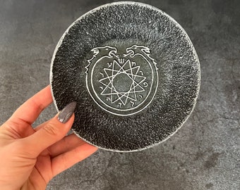 May include: A round, black coaster with a textured surface. The coaster features a white, stylized illustration of two dragons encircling a star-like symbol. The coaster is held by a hand.