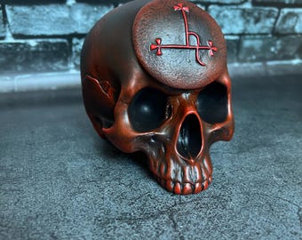 May include: A dark reddish-brown skull sculpture featuring a circular emblem on the forehead. The emblem displays a red linear symbol with cross-like terminals. The skull has deep, dark eye sockets and a detailed jawline with visible teeth.