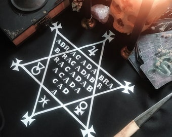 May include: A black cloth with a white pentagram and the words "ABRACADABRA" written around it. There are candles, a skull, a bone, and tarot cards on the cloth.