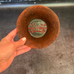 Op de afbeelding: Een ronde, getextureerde, oranje en zwarte keramische kom. De kom heeft een donkergrijze kern met een rood, gestileerd ontwerp. De kom wordt door een persoon vastgehouden.
