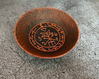 May include: A black bowl with an orange rim and a black and white sigil inside. The sigil is a five-pointed star with a circle around it and other symbols inside the circle.