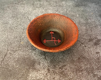 May include: A small, round, shallow bowl with a textured, dark charcoal-gray interior and a vibrant orange-red rim. The center of the bowl features a distinct, bright red sigil or symbol with cross-like terminals, set against the dark base.